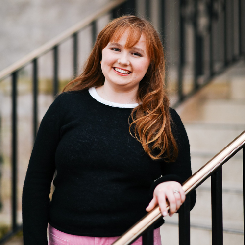 Emma has long red hair, and wears a black sweater while standing on an outdoor staircase.